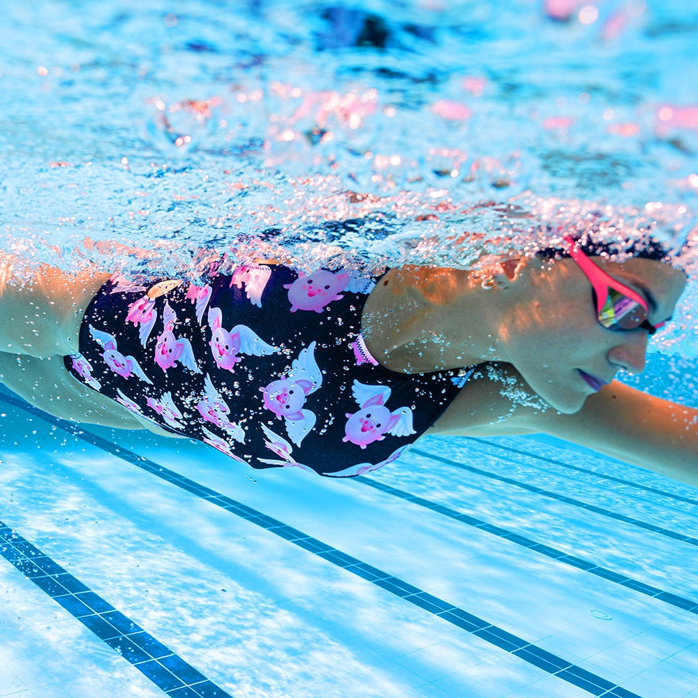 Modella nuota a stile libero con occhialini e testa sott'acqua indossando un costume da allenamento nuoto Boneswimmer, modello intero PINUP 404p con stampa di graziosi e tondi maiali volanti su sfondo nero